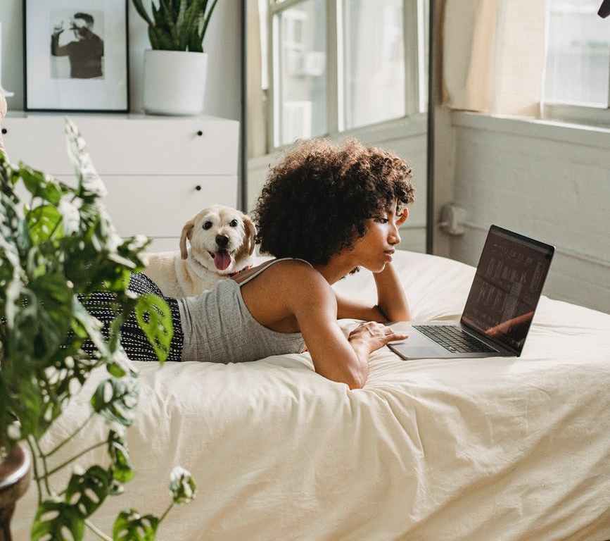 relaxed black woman watching laptop near dog on bed